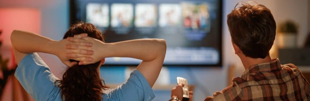 Two people sit on a sofa watching a large smart TV displaying a streaming service interface with rows of content thumbnails. One person holds a remote control while the other leans back with hands behind their head, creating a relaxed home entertainment scene.