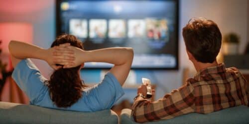 Two people sit on a sofa watching a large smart TV displaying a streaming service interface with rows of content thumbnails. One person holds a remote control while the other leans back with hands behind their head, creating a relaxed home entertainment scene.