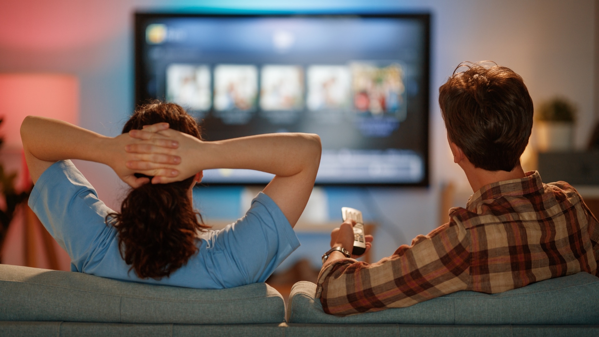 Two people sit on a sofa watching a large smart TV displaying a streaming service interface with rows of content thumbnails. One person holds a remote control while the other leans back with hands behind their head, creating a relaxed home entertainment scene.